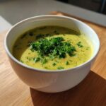 A close-up of a bowl of creamy, yellow-green zucchini soup topped generously with fresh chopped parsley.