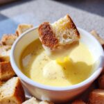 A piece of toasted bread being dipped into a bowl of creamy, yellow cheese fondue recipe, surrounded by more bread cubes.