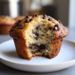 Close-up of a fluffy banana chocolate chip muffin with a bite taken out, revealing melted chocolate inside.