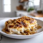 A close-up of a juicy french onion chicken breast topped with melted cheese and caramelized onions on a white plate.