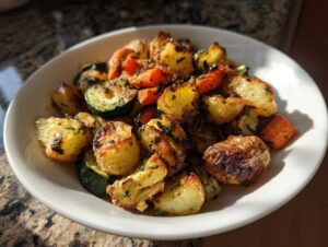A white bowl filled with golden brown Garlic Herb Roasted Veggies, including potatoes, carrots, and zucchini, glistening under sunlight.