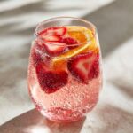 Close-up of a glass filled with bubbly pink champagne punch, garnished with fresh strawberry slices and orange wedges.