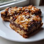 Close-up of two rich magic cookie bars layered with chocolate, toasted coconut, and white coconut flakes on a white plate.