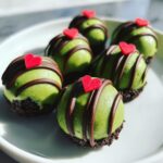 Six bright green grinch oreo balls sitting on a white plate, drizzled with chocolate and topped with small red hearts.