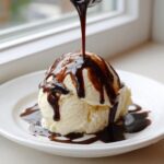 A scoop of vanilla ice cream being drizzled with thick, homemade chocolate syrup from a spoon.