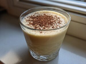 A glass of creamy homemade eggnog topped with a dusting of cocoa or nutmeg, sitting near a window.