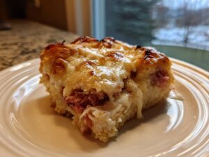 A close-up of a square serving of homemade Reuben bake, featuring melted cheese and corned beef on a white plate.