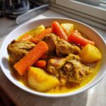 Close-up of a white bowl filled with rich, yellow Jamaican curry chicken pieces, large chunks of carrots, and potatoes.