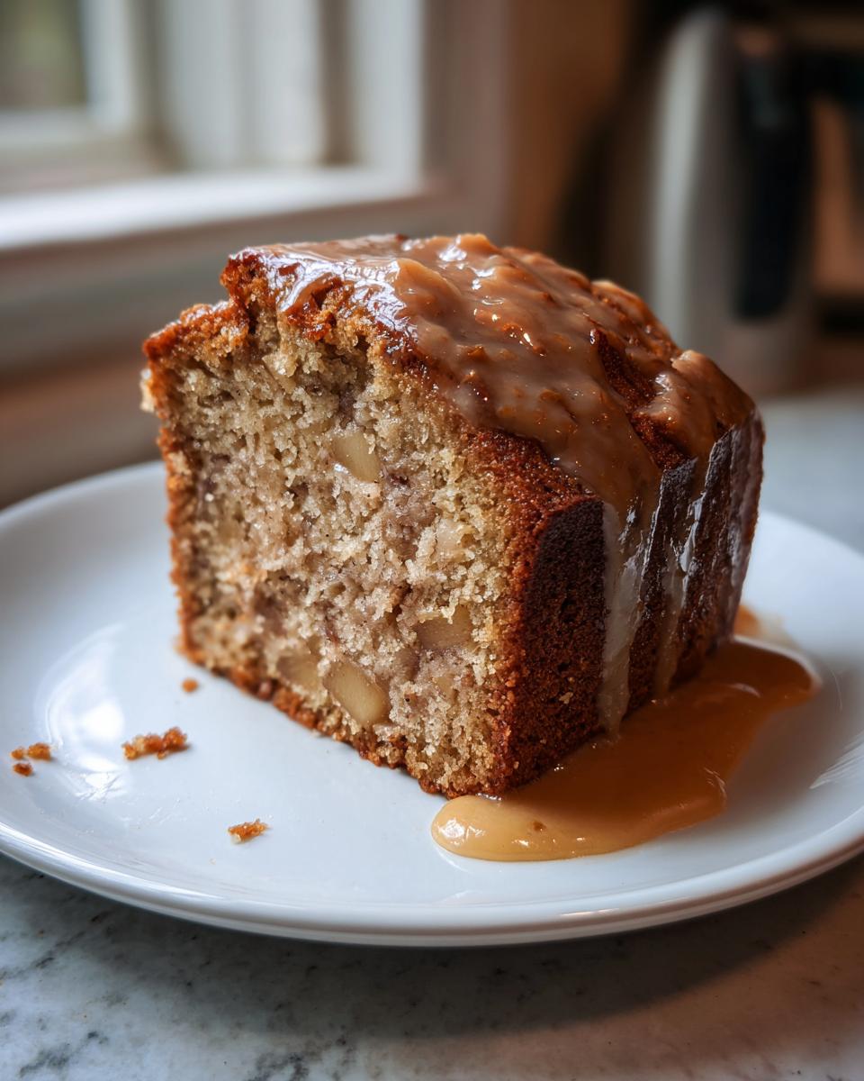 A thick slice of moist apple dapple cake topped with a thick caramel glaze, served on a white plate.