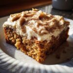 A close-up of a moist slice of applesauce cake topped with thick, swirled frosting and dusted with cinnamon.