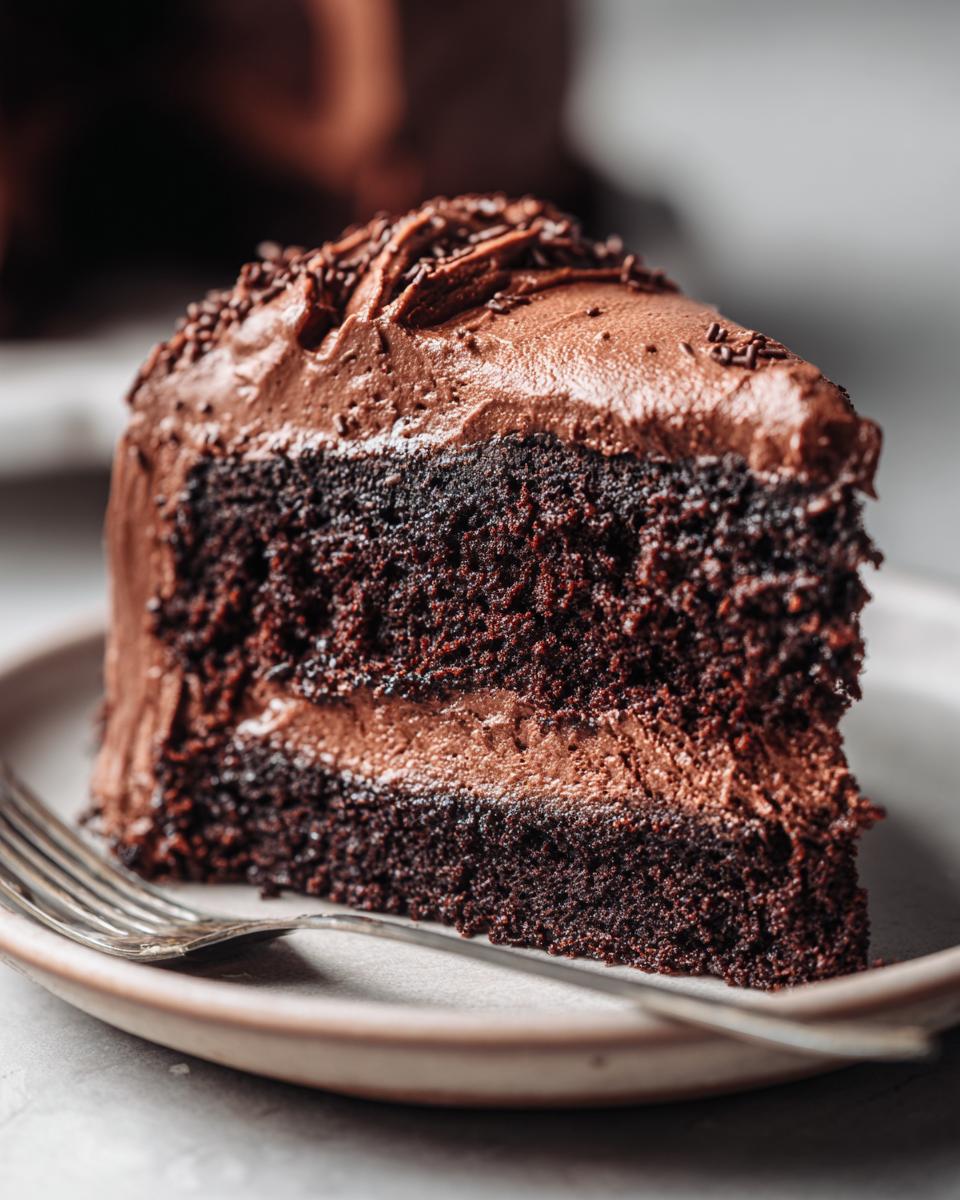 Close-up of a rich, two-layer slice of chocolate cake topped with thick chocolate frosting and sprinkles.