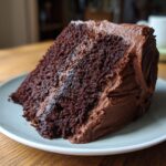 A close-up of a generous slice of moist chocolate cake covered in thick chocolate frosting.