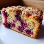 Close-up of a moist slice of plum cake showing purple fruit baked into the yellow crumb and a brown streusel topping.