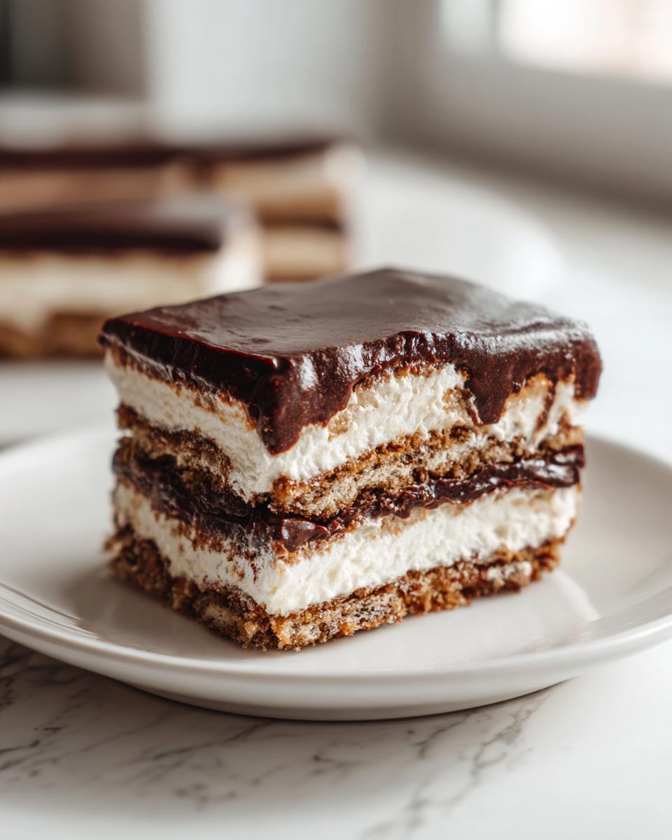 A square slice of no-bake eclair cake featuring layers of graham crackers, white cream filling, and topped with shiny chocolate ganache.