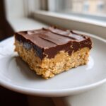 A close-up of a single, thick no-bake peanut butter bars square with a chocolate topping, resting on a white plate.