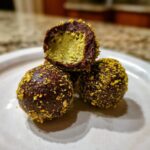 Close-up of three decadent dubai chocolate balls coated in crushed pistachios, one cut open revealing a bright green filling.