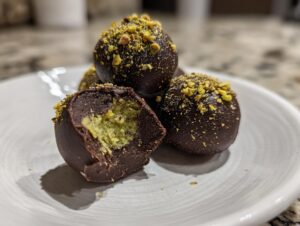 Close-up of rich, dark chocolate dubai chocolate balls topped with crushed pistachios, one cut open showing a bright green filling.