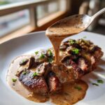 A spoonful of rich, creamy mushroom sauce is being poured over sliced steak topped with sautéed mushrooms.