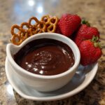 A small white bowl filled with glossy, melted chocolate dip, served alongside fresh strawberries and pretzels.