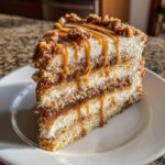 A tall slice of caramel crunch cake featuring multiple layers of cake, cream, and crunchy topping, drizzled with caramel sauce.