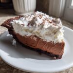 A generous slice of french silk pie featuring a dark chocolate crust, rich chocolate mousse filling, and topped with whipped cream and cocoa powder.