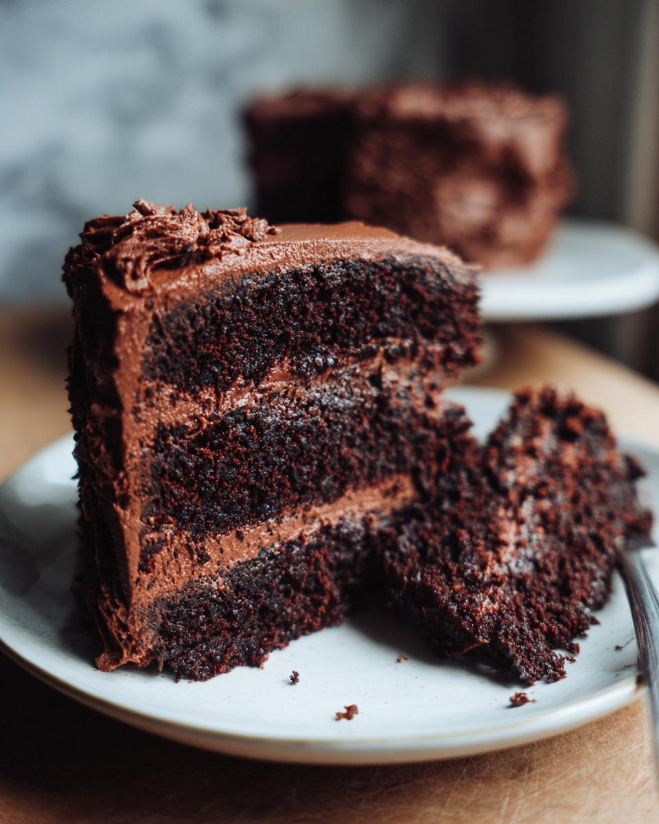 A rich, moist slice of three-layer chocolate cake with chocolate frosting, served on a plate.