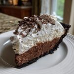 A decadent slice of chocolate mud pie featuring a dark crust, rich chocolate filling, and thick whipped cream topped with chocolate shavings.
