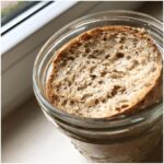 Close-up of a slice of bread resting on top of a bubbly sourdough starter inside a glass jar.