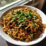 A close-up of a white bowl filled with glossy, savory fried noodles, topped generously with fresh green onions.
