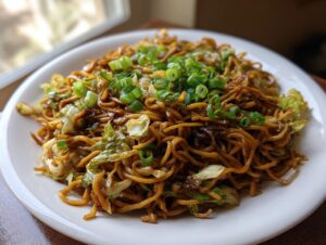 A plate of glossy, dark brown fried noodles mixed with wilted cabbage and topped with fresh green scallions.