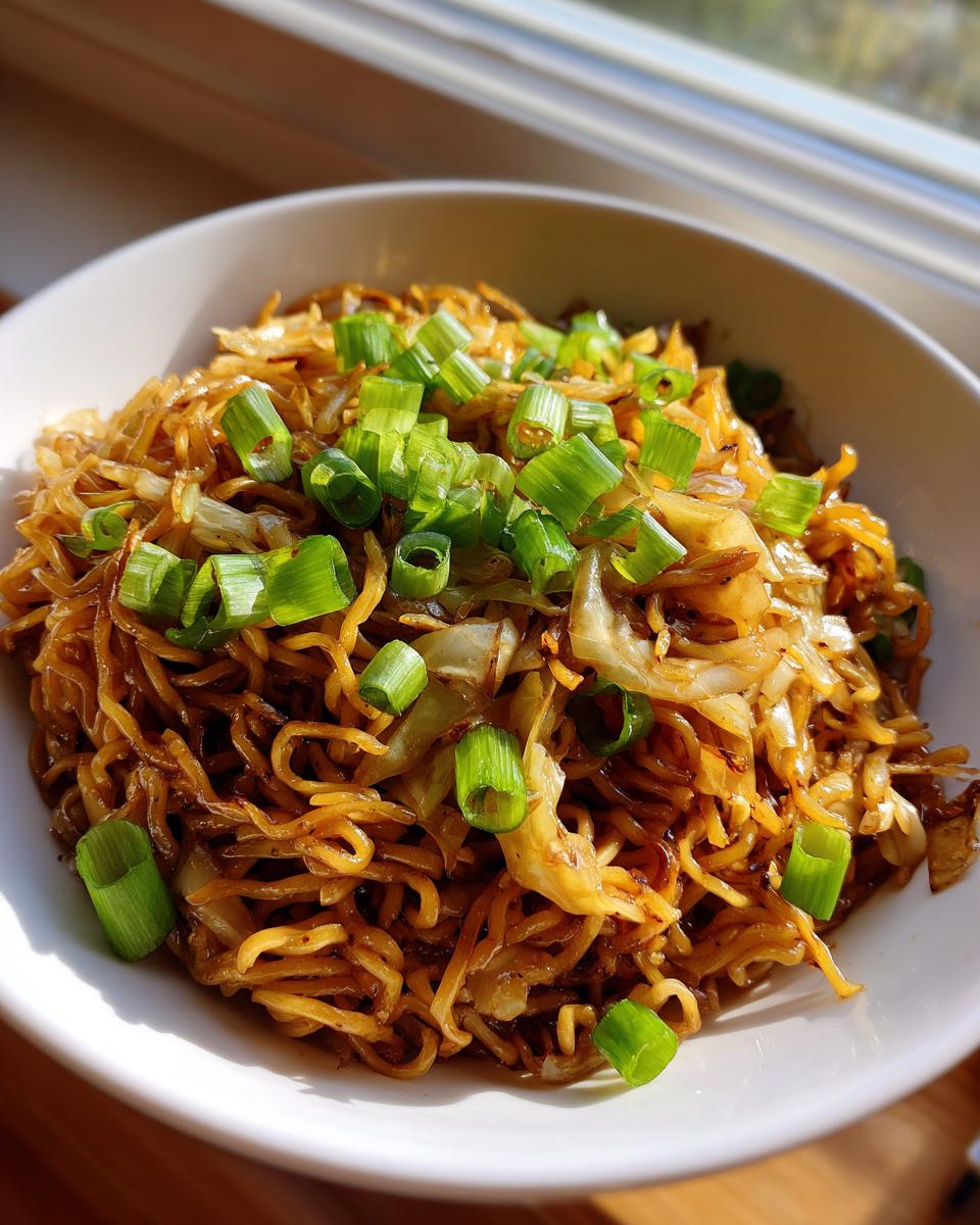 A close-up of a white bowl filled with glossy, brown fried noodles and sautéed cabbage, topped generously with bright green chopped scallions.
