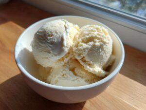 Three scoops of creamy vanilla protein ice cream served in a white bowl on a wooden surface.