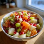 Close-up of a vibrant winter fruit salad featuring mandarin oranges, kiwi, pomegranate seeds, and grapes in a white bowl.