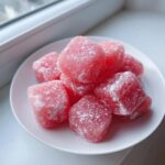 A close-up of several pink, cube-shaped watermelon mochi candies dusted with white powdered sugar on a small white plate.
